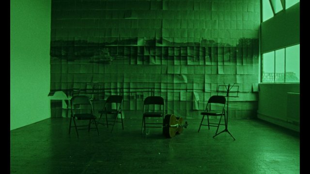 Video Reference: empty room, folding chairs, music stands, guitar, interior, green lighting, stage setup, rehearsal space, window, abstract wall, floor, minimal, indoor