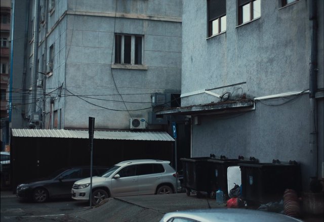 Video Reference: alley, urban, apartment building, parked cars, trash bins, overhead wires, air conditioner, narrow street, gray exterior, residential area, parking, service lane, building facade, outdoor, city