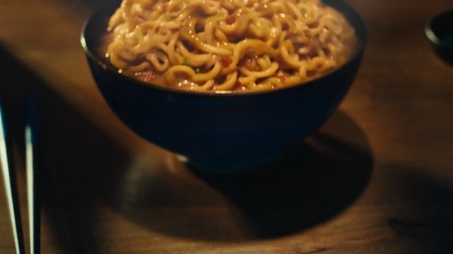 Video Reference: noodles, bowl, food, steaming, meal, close-up, dinner, wooden table, warm light, indoor, cooked noodles, soup, comfort food