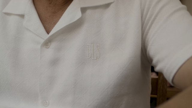 Video Reference: close-up, shirt, white clothing, button-up, collar, embroidered, monogram, torso, fabric, casual, indoor, person