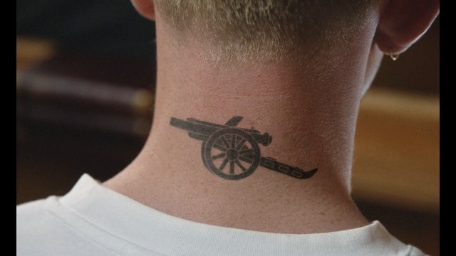 Video Reference: neck tattoo, tattoo, close-up, back of head, white shirt, cannon wheel, emblem, person, indoors, blurred background