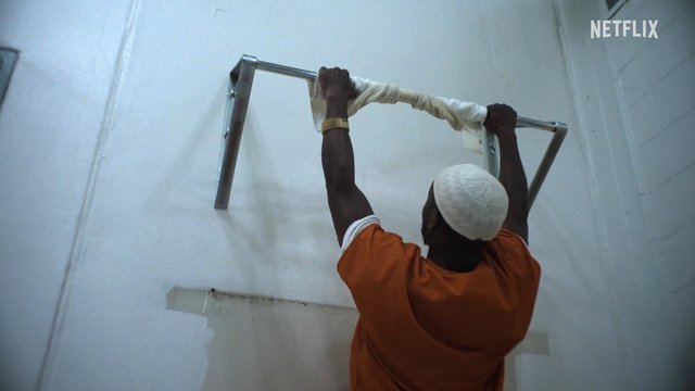 Video Reference: person, man, orange shirt, white cap, wall bar, exercise, indoor, interior, reaching, pullup, fitness, plain wall, metal bar, low angle, Netflix, worn cloth