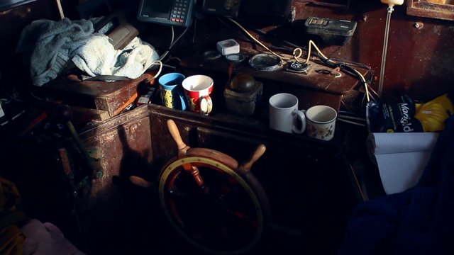Video Reference: cluttered interior, wooden helm, steering wheel, mugs, towels, cables, electronic devices, small cabin, boat cabin, dim light, messy room, personal items, wooden surfaces, notebook, controls, storage