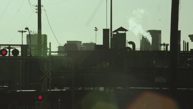 Video Reference: industrial, city skyline, smoke, steam, silhouette, utility poles, railroad crossing, traffic lights, rooftop, urban, buildings, pipes, sunlight, haze