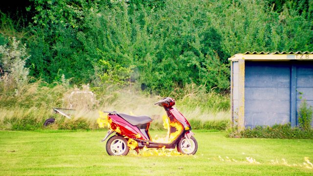 Video Reference: scooter, fire, burning, flames, grass, outdoors, vehicle, red, smoke, shed, greenery, rural, accident, damage