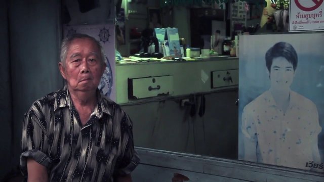 Video Reference: elderly man, indoor, portrait poster, shop, counter, seated, interior, street scene, serious expression, window display, Thai text, barbershop, small business, low light