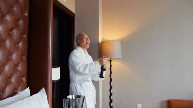 Video Reference: hotel room, man, robe, champagne, celebration, indoor, lamp, bedroom, older adult, smiling, luxury, casual, interior, bottle, relaxation