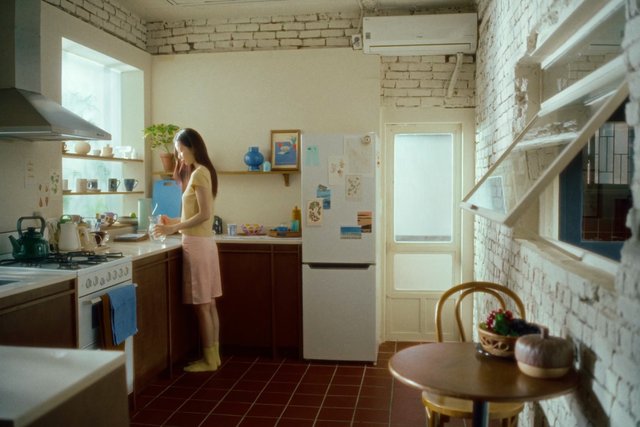 Video Reference: kitchen, woman, indoor, home, cooking, counter, refrigerator, stove, appliances, sunlight, interior, domestic, table, shelves, brick wall, apartment