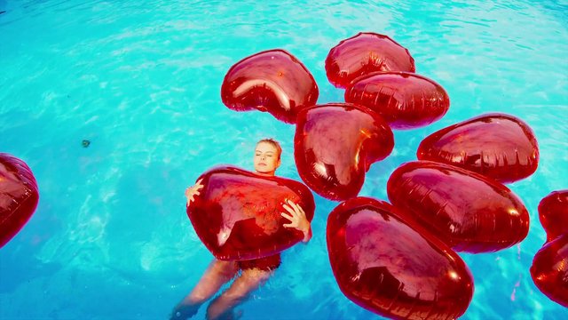 Video Reference: swimming pool, floating, heart balloons, red balloons, water, person, inflatable, summer, blue water, recreation, overhead view, surreal