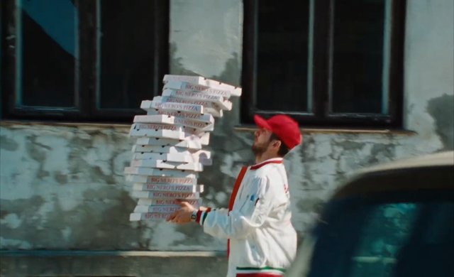 Video Reference: man, pizza delivery, pizza boxes, stack, red cap, uniform, outdoors, street, building, side view, delivery, motion blur, urban, food service