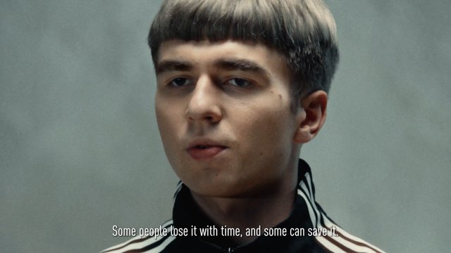 Video Reference: young man, close-up, portrait, track jacket, striped collar, gray background, subtitle, indoor, serious expression, bowl haircut