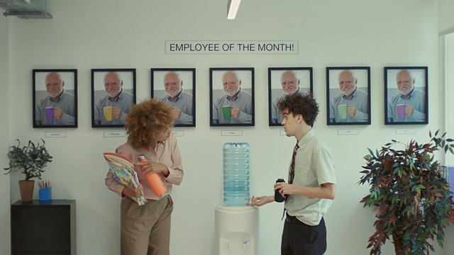 Video Reference: office, break room, water cooler, employee of the month, framed photos, two people, conversation, indoor, plants, workplace, wall art, standing, casual office