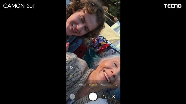 Video Reference: smartphone, camera app, selfie, two people, smiling, outdoors, portrait mode, interface, screen capture, mobile photography, close-up, casual, daylight, brand text